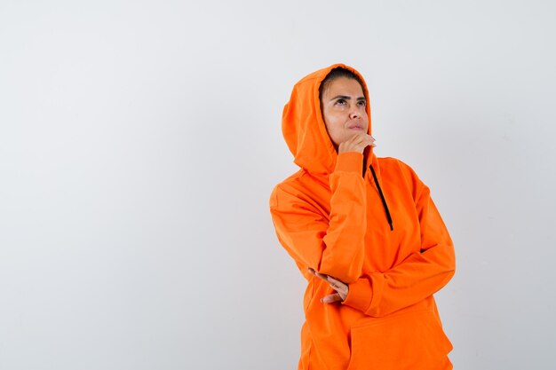 Женщина в оранжевой толстовке с капюшоном подпирает подбородок рукой и выглядит задумчивой