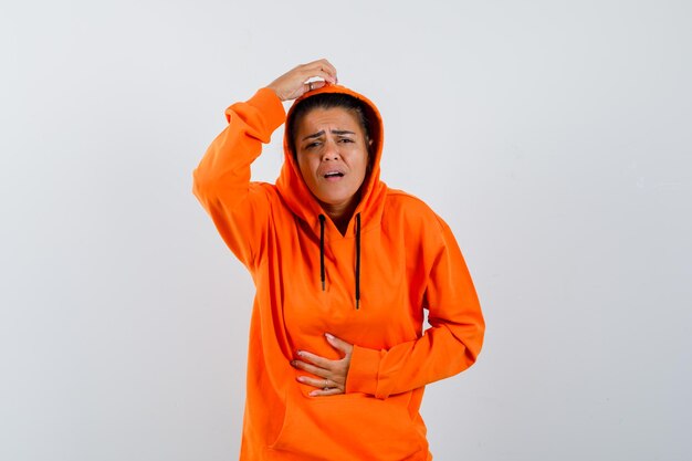 Женщина в оранжевой толстовке с капюшоном страдает от боли в животе и плохо выглядит