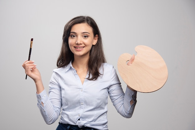 Foto gratuita pittore femminile in posa con tavolozza e pennello sul muro grigio.