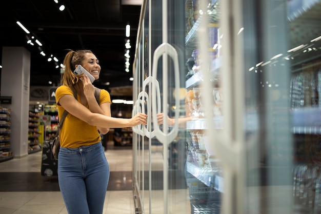 Female person with shopping cart opening fridge to take food in grocery store while talking on the phone