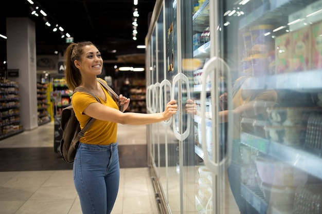 Female person with shopping cart opening fridge to take food in grocery store