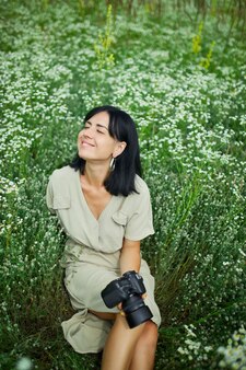 카메라를 들고 있는 여성 사진작가는 야외에서 눈을 감고 휴식을 취하고 있는 꽃밭에 앉아 휴식을 취하고 디지털 카메라를 손에 들고 있습니다. 여행 자연 사진, 텍스트를 위한 공간, 평면도.