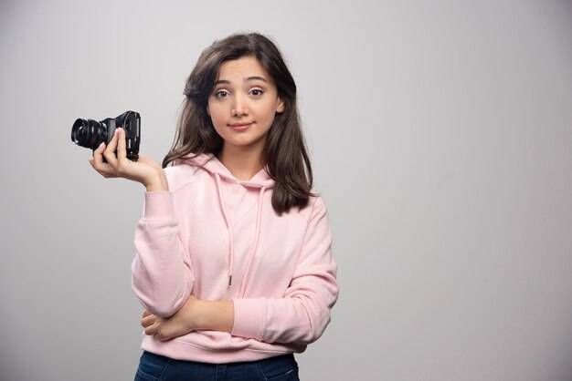 Female photographer posing with camera on gray wall.