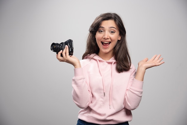 Female photographer posing with camera on gray wall. 