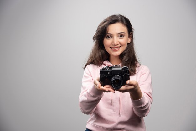 Женский фотограф улыбается с камерой на серой стене.