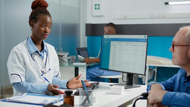 Female physician consulting elderly man with physical impairment, having medical appointment in office. Doctor talking to patient wheelchair user with disability, giving healthcare support.