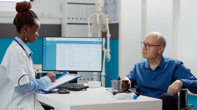 Medico femminile che prende ntoes alla visita di controllo con l'utente su sedia a rotelle, facendo consulenza in studio medico. medico che esamina il paziente anziano con disabilità cronica e menomazione.