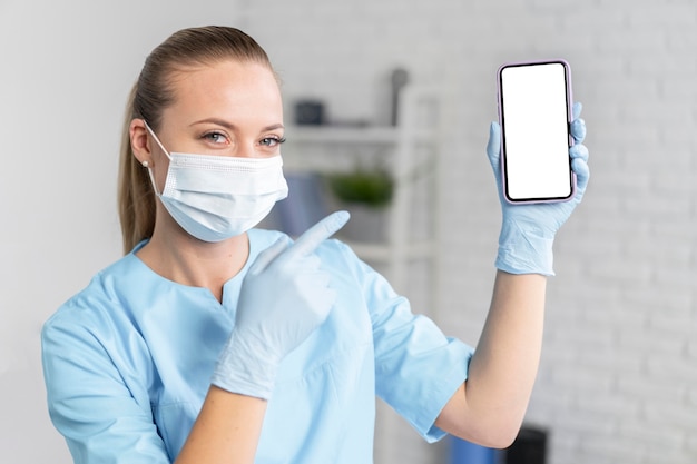 Бесплатное фото Женский физиотерапевт с медицинской маской, держащей и указывающей на смартфон