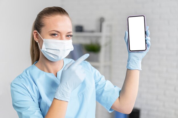 Female physiotherapist with medical mask holding and pointing at smartphone