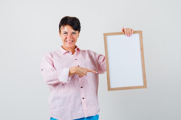 Donna in camicia rosa, pantaloni che punta alla cornice vuota e che sembra allegra, vista frontale.