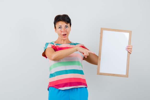 Female pointing at empty frame in striped t-shirt, pants and looking amazed. front view.