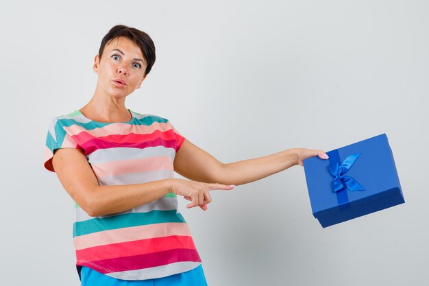 Female pointing at present box in striped t-shirt, pants and looking surprised , front view.