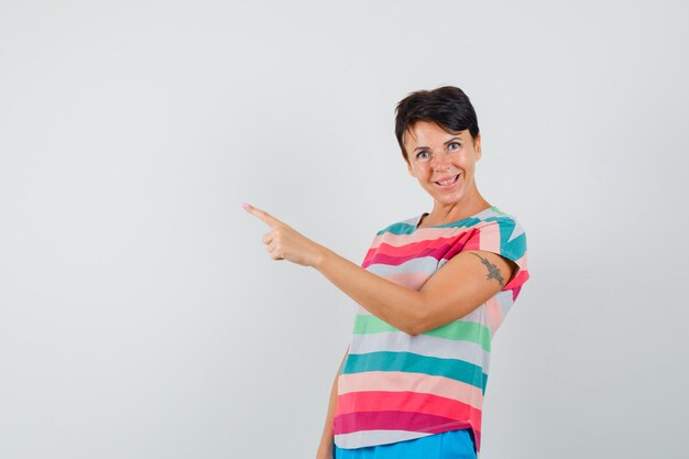 Female pointing at upper left corner in striped t-shirt, pants and looking merry. front view.