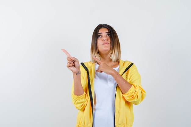 Female pointing at upper left corner in t-shirt, jacket and looking hesitant , front view.