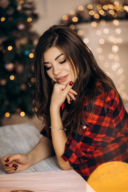 Female portrait. Pregnant woman in checked shirt poses in cozy room with Christmas tree