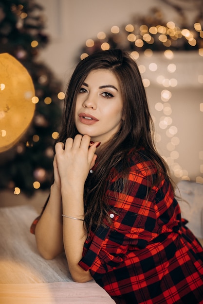 Female portrait. Pregnant woman in checked shirt poses in cozy room with Christmas tree