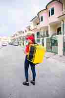 Foto gratuita postino femminile in berretto rosso in piedi con borsa termica gialla e tenendo la compressa. deliverywoman che consegna l'ordine a piedi.