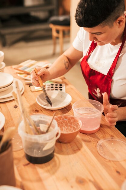 Female potter painting ceramic dishware with paintbrush in workshop