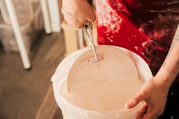 Рука женского гончара вставляет тарелку в ведро с краской