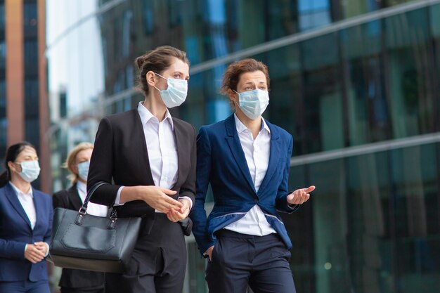 Female professionals wearing office suits and masks, meeting and walking together in city, talking, discussing project. Medium shot. Pandemic and business concept