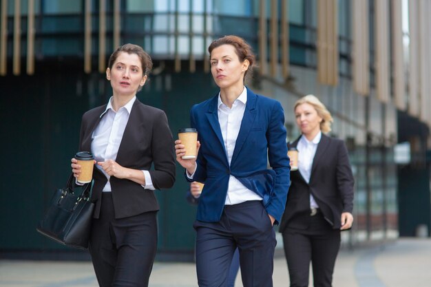 Female professionals with paper coffee cups wearing office suits, walking together in city, talking, discussing project or chatting. Front view. Businesswomen outdoors concept