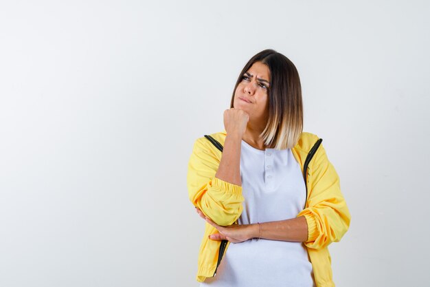 Female propping chin on fist in t-shirt, jacket and looking thoughtful , front view.