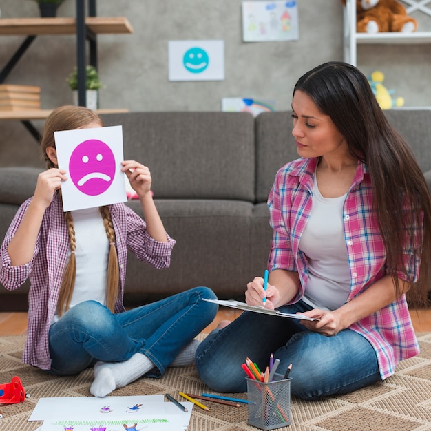 Female psychologist taking notes looking at girl covering her face with sad emotion card