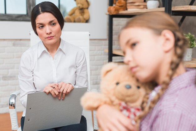 Female psychologist with clipboard and pen observing the depressed girl hugging her teddy bear