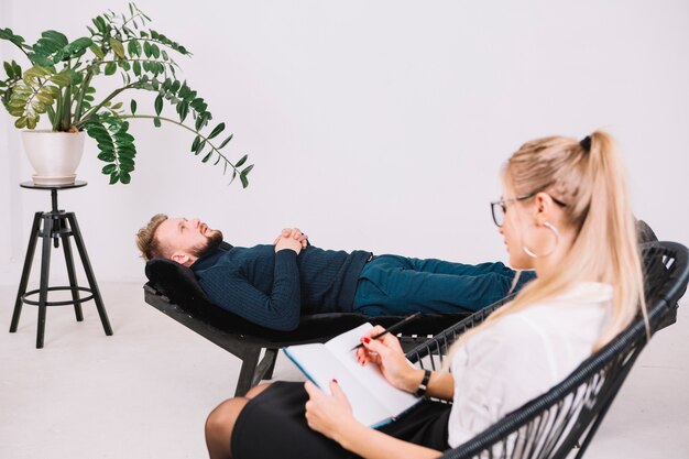 Female psychologist writing notes on diary while patient lying on couch in clinic