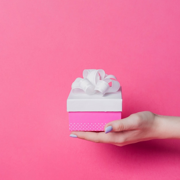 Female's hand holding box with white ribbon bow on pink background