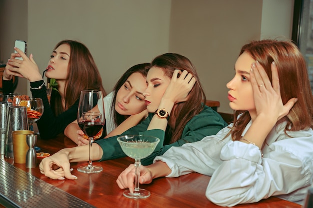 Foto gratuita amici femminili tristi e stanchi che bevono un drink al bar. sono seduti a un tavolo di legno con cocktail. indossano abiti casual.