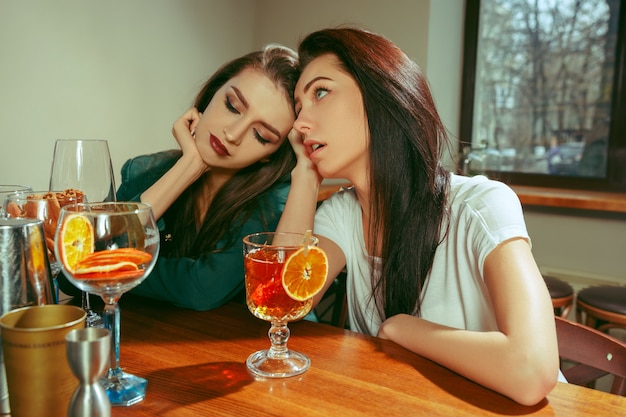 Amici femminili tristi e stanchi che bevono un drink al bar. sono seduti a un tavolo di legno con cocktail. indossano abiti casual.
