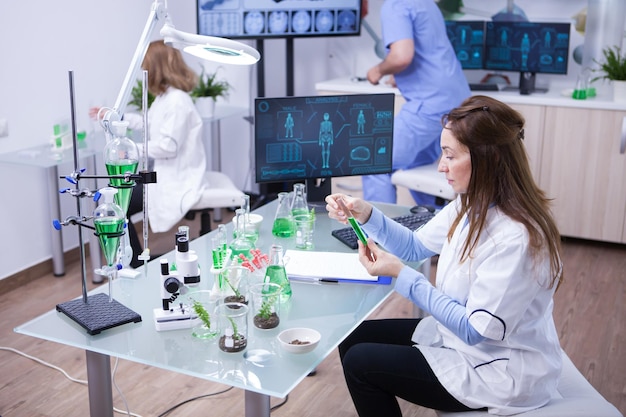 Scienziata in possesso di una soluzione verde mentre ascolta il team lavora in background. sullo sfondo un giovane biologo.