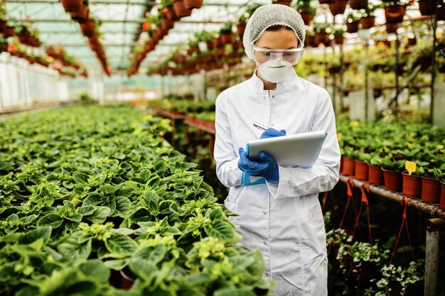 鉢植えの花の成長を検査し、温室でメモを取る女性科学者