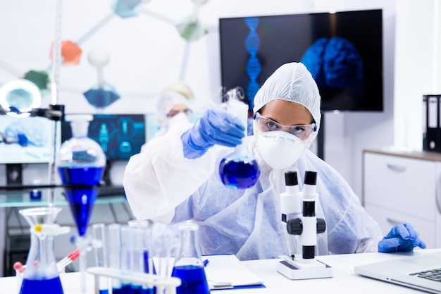 파란색 솔루션이 있는 시험관을 들고 보고 있는 보호 장비를 입은 여성 과학자. 화학 실험실입니다.
