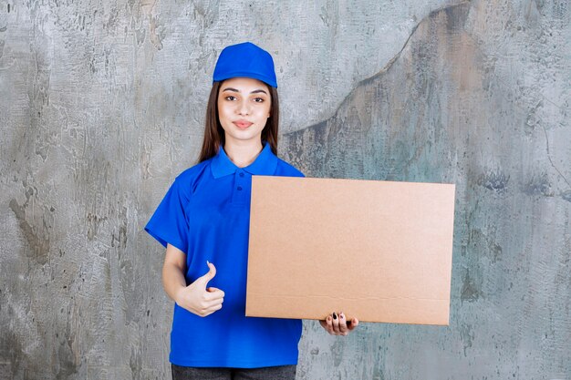 Female service agent in blue uniform holding a cardboard box and showing positive hand sign.