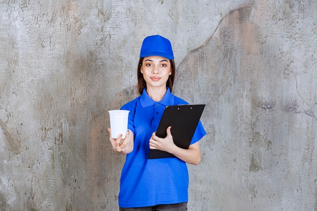 Foto gratuita agente di servizio femminile in uniforme blu che tiene una tazza monouso bianca e una cartella cliente nera.