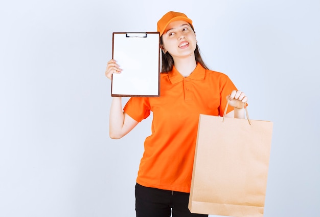 Female service agent in orange color dresscode holding a cardboard shopping bag and presenting the signature list to the customer while looking thoughtful.