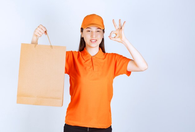 Female service agent in orange color dresscode holding a cardboard shopping bag and showing successful hand sign meaning quality assurance