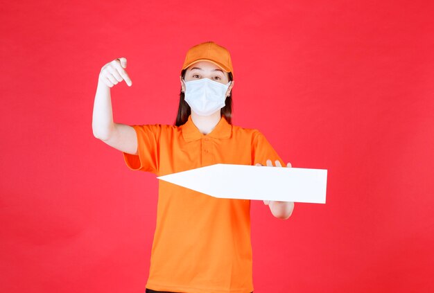 Female service agent in orange color dresscode and mask holding an arrow pointing to the left