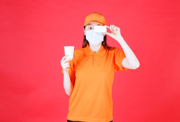 Female service agent in orange color dresscode and mask holding a disposable cup and presenting her business card