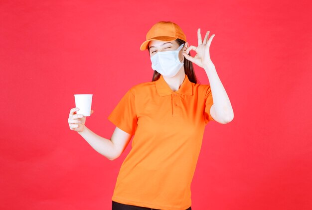 Агент женской службы в оранжевом дресс-коде и маске держит одноразовую чашку и показывает знак рукой