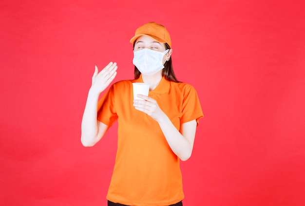 Female service agent in orange color dresscode and mask holding a disposable cup and smelling the product.