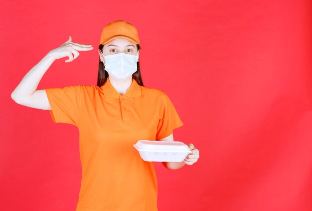 Female service agent in orange color dresscode and mask holding a takeaway food package and looks thoughtful and dreaming