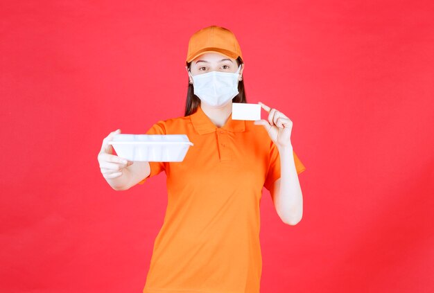 Female service agent in orange color dresscode and mask holding a takeaway food package and presenting her business card