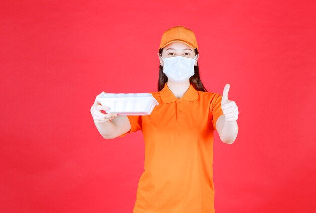 Female service agent in orange color dresscode and mask holding a takeaway food package and showing positive hand sign