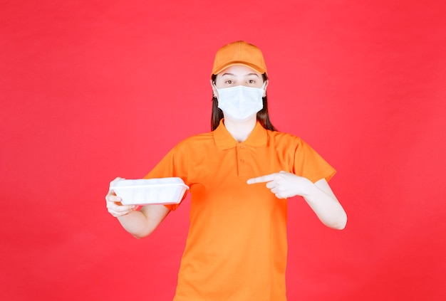 Female service agent in orange color dresscode and mask holding a takeaway food package