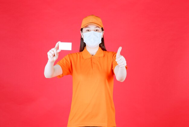 Female service agent in orange color dresscode and mask presenting her business card and showing positive hand sign