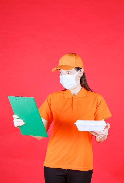 주황색 유니폼과 테이크 아웃 식품 패키지를 들고 녹색 폴더를 확인하는 마스크의 여성 서비스 요원.
