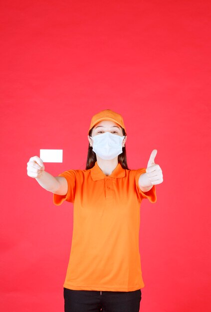 Female service agent in orange color uniform and mask presenting her business card and showing positive hand sign.